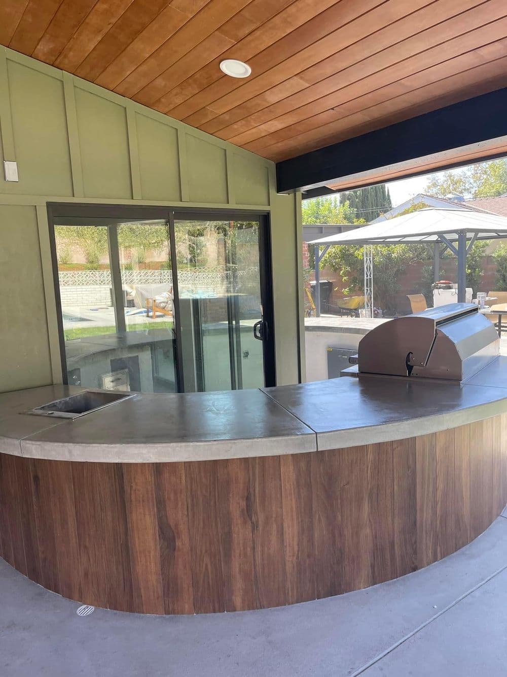 Modern outdoor kitchen with curved concrete countertop and built-in grill, surrounded by glass walls.