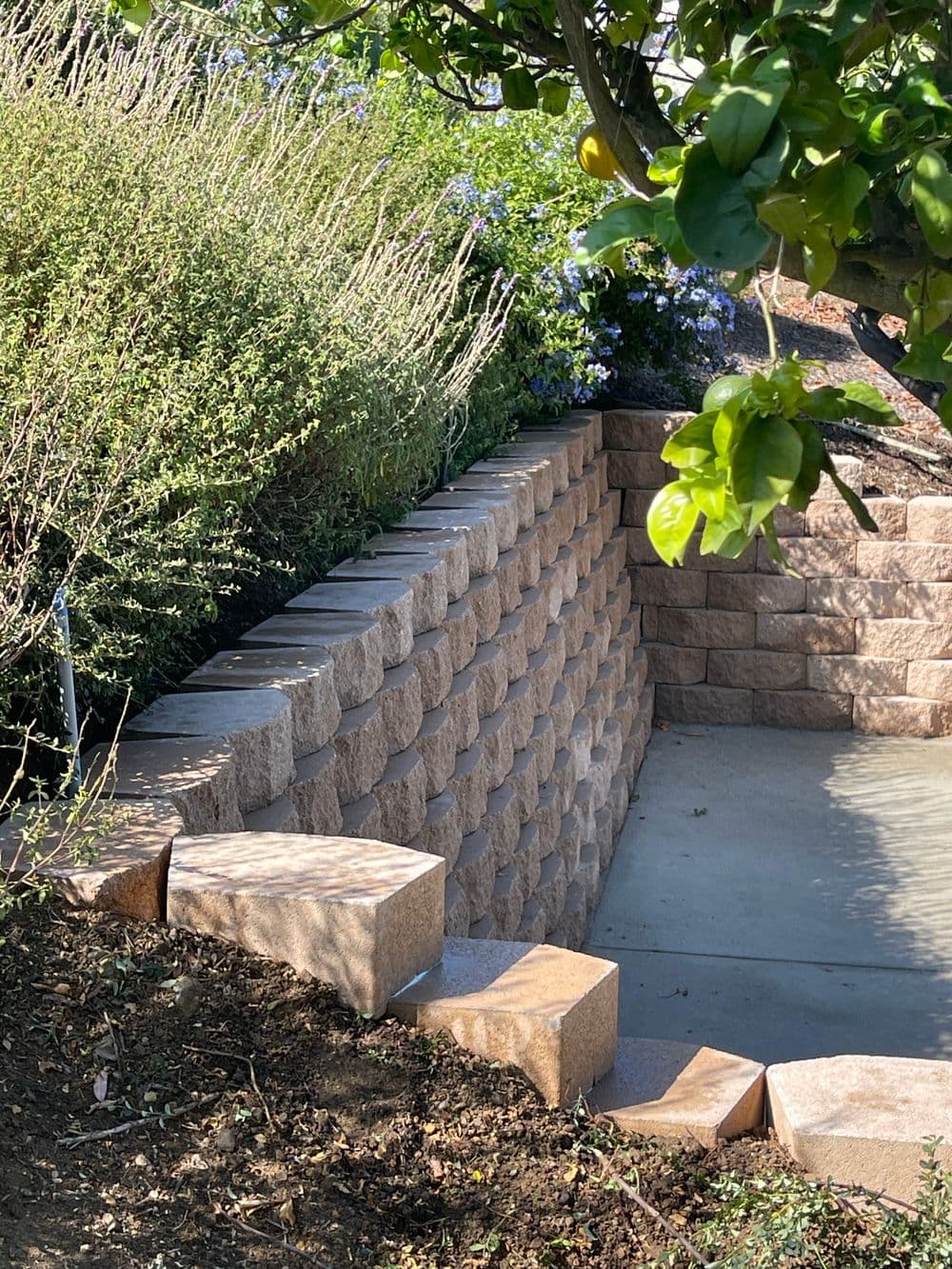 Stone retaining wall with steps, surrounded by greenery and flowers in a garden setting.