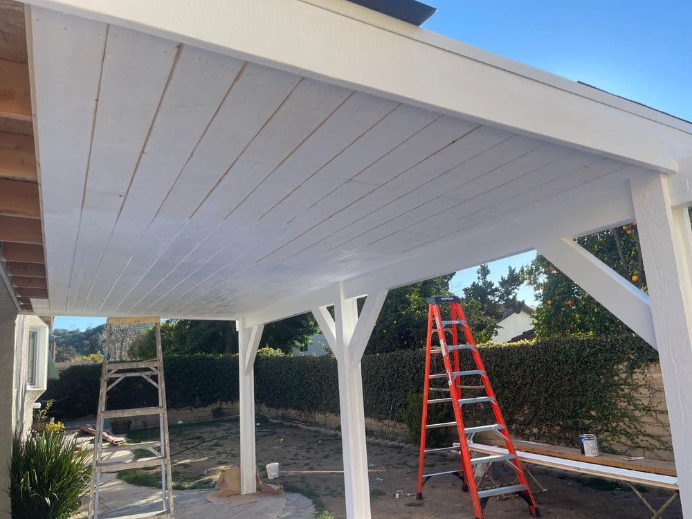 White wooden patio ceiling with a ladder and scaffolding in a sunny backyard setting.