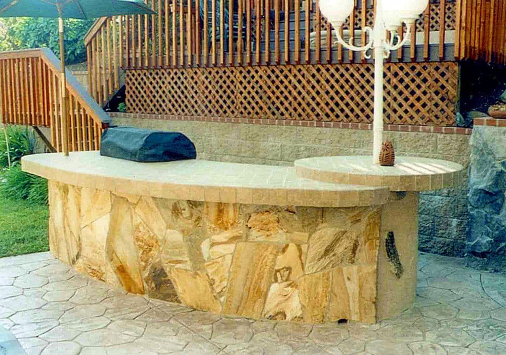 Outdoor bar with a curved stone countertop and grill, set in a backyard with wooden features.